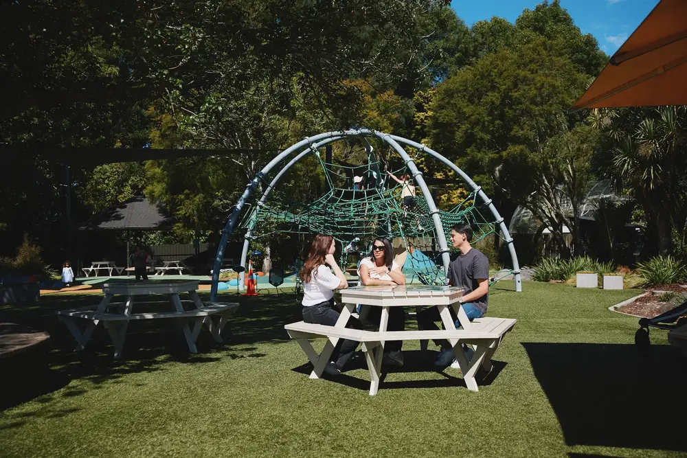 A group of adults sitting at a picnic table in front of the playground at Hamilton Zoo